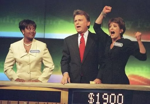 Johnna Goodwin, left, watches as Donna Handel, right, reacts after winning on "Wheel of Fortune," with host Pat Sajak during a taping of the game show in Philadelphia on April 17, 1999. The game show was in Philadelphia for the weekend taping a week's worth of shows. Ryan Seacrest will replace the retiring Sajak as host of “Wheel of Fortune.” (AP Photo/William Thomas Cain, File)