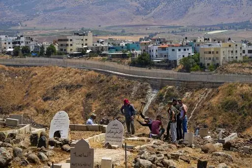 Residents dig a grave at a cemetery on the Lebanese side of the Lebanese-Israeli border in the southern village of Wazzani with the walled northern part of Ghajar village in the background, Lebanon, Tuesday, July 11, 2023. The little village of Ghajar has been a point of contention between Israel and Lebanon for years, split in two by the border between Lebanon and the Israeli-occupied Golan Heights. The dispute has begun to heat up again.(AP Photo/Hassan Ammar)