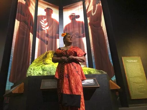 Hinaleimoana Wong-Kalu, one of the curators of the new Kapaemahu exhibit at Bishop Museum, poses for a photo in Honolulu on Thursday, June 16, 2022 in front of pictures of four healers who visited Hawaii from Tahiti more than 500 years ago. The exhibit draws attention to the stories of the healers, who were "mahu" or individuals who presented themselves as a mixture of male and female, and highlights gender fluidity's deep roots in Polynesia. (AP Photo/Audrey McAvoy)