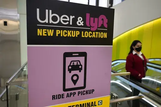 A passerby walks past a sign offering directions to an Uber and Lyft ride pickup location at an airport, Feb. 9, 2021, in Boston. Drivers for Uber and Lyft will earn a minimum pay standard of $32.50 per hour under a settlement announced, Thursday, June 27, 2024, by Massachusetts Attorney General Andrea Campbell, a deal that also includes a suite of benefits and protections. (AP Photo/Steven Senne, File)