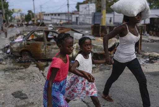 Girls holding hands are led past a burnt car blocking the street as they evacuate the Delmas 22 neighborhood to escape gang violence in Port-au-Prince, Haiti, May 2, 2024. As young Haitians are increasingly exposed to violence, the country is undergoing a wider push to dispel a long-standing taboo on seeking therapy and talking about mental health. (AP Photo/Ramon Espinosa, File)